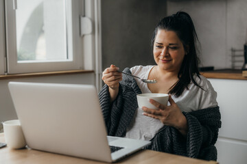Overweight woman eats oatmeal and looks at laptop