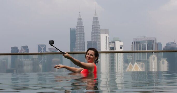 Woman on long-awaited vacation after year of work bathes in pool on roof of skyscraper. Happy woman records video with small camera and selfie stick, relax and enjoy life in open air swimming pool