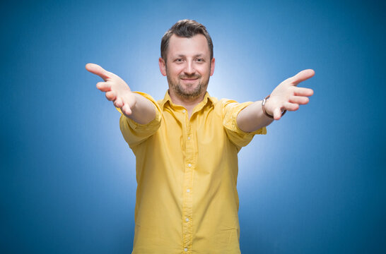Half length lovely man with open arms going to you over blue background, dresses in yellow shirt. Welcome concept