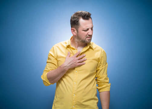 Half length man suffering from breathing problem over blue background, dresses in yellow shirt. Coughing, guy in pain touching chest respiratory symptoms fever