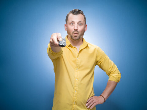 Amazed man holding remote control watching TV - movies over blue background, dresses in yellow shirt. Its weekend. Front view of surprised guy shows television