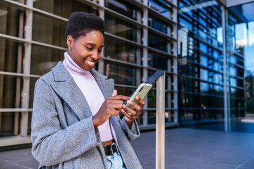 Young African American female in stylish coat and sweater smiling and browsing news on cellphone. She is leaning on railing outside modern building on city street