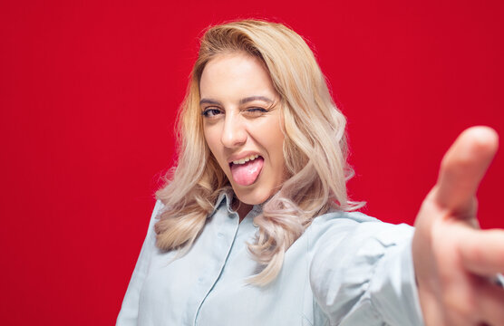 Selfie. Close-up funny mocked woman takes photo of himself with her smartphone while tongue sticking out, isolated on red background. Focus on face