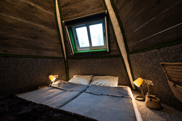 Double bed in bedroom of pyramid shape wooden cabin. Pyramid shape walls of bungalow from inside. Small room, angled walls, window light, ceiling lamp, tree trunks corners, made bed.