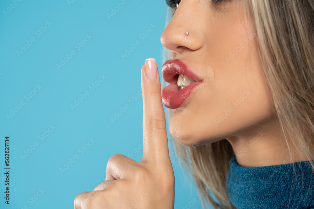 Wall mural Young woman with finger over her mouth, silence gesture on a blue background, close up