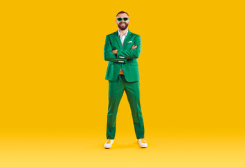 Portrait of cheerful cool and joyful man in green suit and sunglasses of same color. Funny man who celebrates St. Patrick's Day standing with folded hands and smiling at camera on orange background.