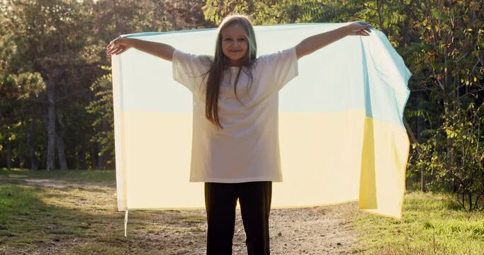 A young beautiful girl unfolds a large flag of Ukraine with her hands in the park in the rays of the evening sun. Wraps herself in the flag of Ukraine and smiles.