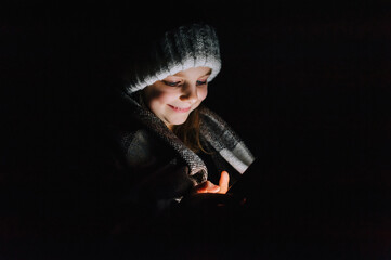 Blackout in Ukraine. Ukrainian beautiful girl, a child in warm clothes, a hat, a blanket, sits in a dark, cold room, looks at a smartphone, phone and plays games.