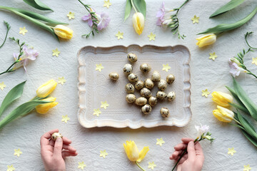 Natural Easter decor, overhead, flat lay with quail eggs, feather wreath, yellow tulip flowers on off white textile background. Flat lay, top view low impact springtime background with human hands.