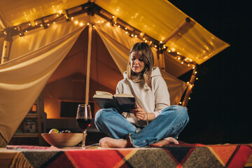 Smiling Woman freelancer drinking wine and read book sitting in cozy glamping tent in autumn evening. Luxury camping tent for outdoor holiday and vacation. Lifestyle concept