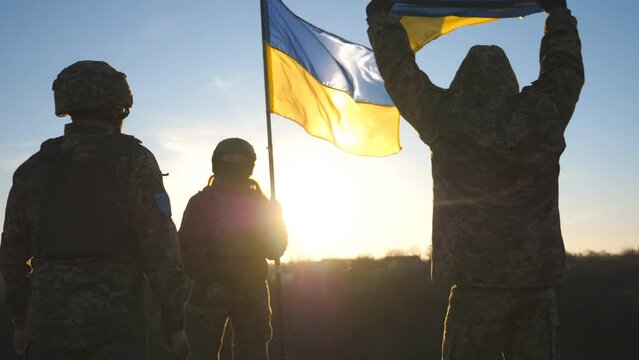 Soldiers of ukrainian army standing at top of hill and lifted up flag of Ukraine. People in military uniform holding waving flag as symbol of victory against russian aggression. End of war in Europe