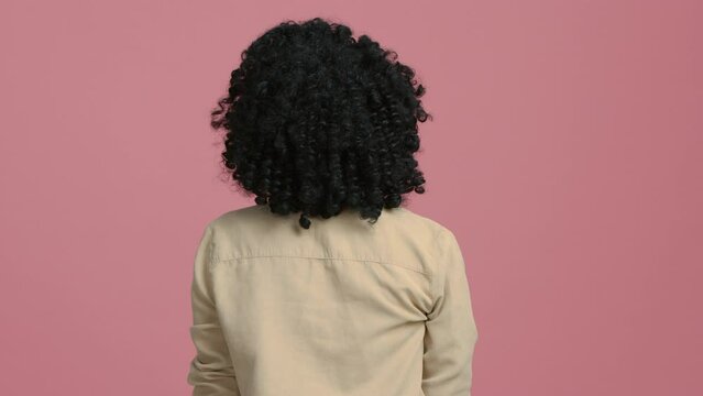 Serious woman of color with afro haircut looking at camera, taking picture on film camera. African American photographer female 20s holding analog camera and taking pictures on pastel pink background