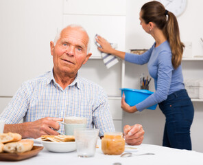 Elderly man drinkimg tea, daughter washing furniture kitchen at home interior