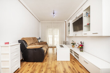 Living room of a house with brown fabric and black skai armchairs and white bookcase with glass shelves and flat tv hanging in the middle