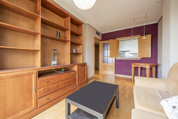 Living room of a house with a fabric armchair, a cherry-colored wooden bookcase and a kitchen with a serving hatch