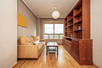 Small living room furnished with a reddish-colored wooden bookcase, a mustard-colored three-seater sofa, and a window with views