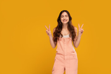 Positive, young, happy woman having fun in studio, showing peace sign, victory gesture and smiling to the camera. Yellow background. Copy space.