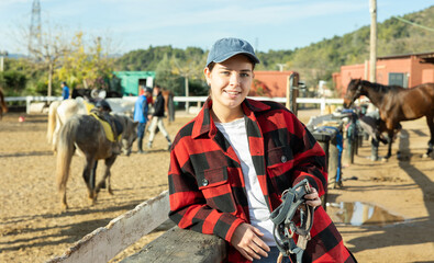 Portrait of positive young female stable worker holding horse halter or harness near wooden fence...