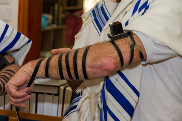 An adult man putting a Jewish Tefillin on his arm and wearing prayer shawl for praying. Preparing...