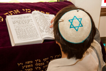 A rear view of praying young man pointing at a phrase in a bible book (sefer torah), while reading a pray - obrazy, fototapety, plakaty
