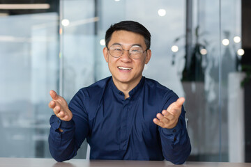 A successful Asian man inside the office is looking at the web camera, talking on a video call with colleagues, gesturing and smiling an online meeting with partners, talking to customers, pov - obrazy, fototapety, plakaty