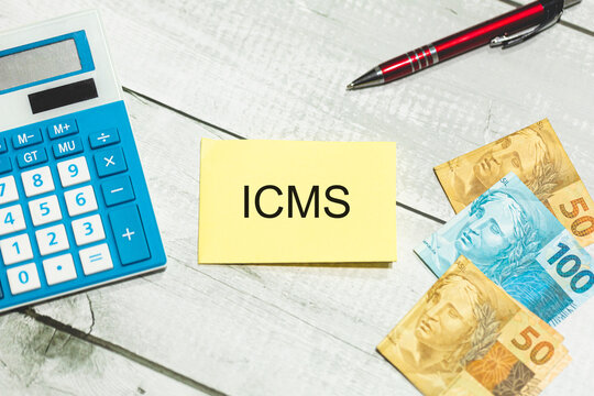 A sigla ICMS referente ao Imposto sobre Circula&ccedil;&atilde;o de Mercadorias e Servi&ccedil;os escrita em um peda&ccedil;o de papel. Notas do Real Brasileiro sobre uma mesa de madeira. Economia Brasileira.
