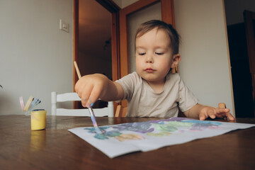 child draws with a brush and multicolored gouache sitting at the table drawing to his mother
