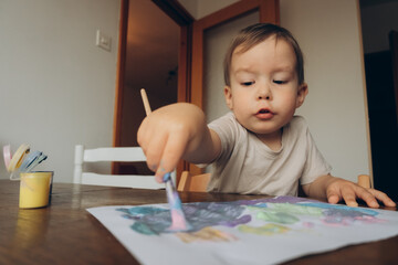 child draws with a brush and multicolored gouache sitting at the table drawing to his mother