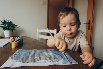 child draws with a brush and multicolored gouache sitting at the table drawing to his mother