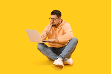 Handsome man with laptop biting nails on yellow background