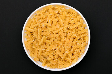 Uncooked Fusilli Pasta Lying on White Plate on Black Background. Raw and Dry Macaroni. Unhealthy and Fat Food - Top View