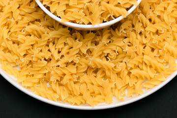 Uncooked Fusilli Pasta Lying on White Plate on Black Background. Raw and Dry Macaroni. Unhealthy and Fat Food - Top View