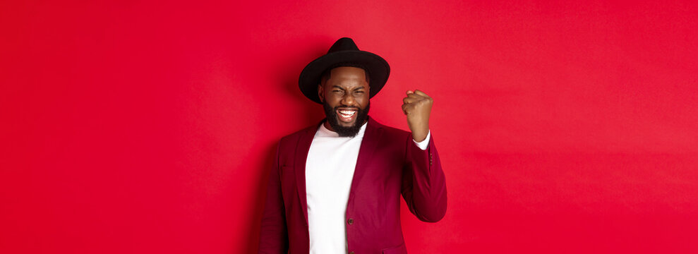 Fashion and party concept. Excited Black man rejoicing of winning, making fist pump gesture and say yes with satisfaction, standing against red background