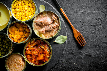 Various open tin cans of canned food on a stone Board with a fork.