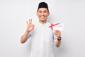 cheerful young Asian Muslim man holding gift certificate coupon voucher, showing okay gesture on...
