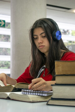 Chica universitaria de blusa roja escribiendo y estudiando en la biblioteca	mientras escucha m&uacute;sica con sus aud&iacute;fonos 