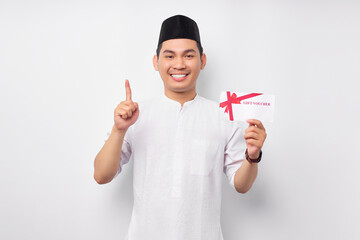 Smiling young Asian Muslim man holding gift certificate voucher, pointing his finger upward...