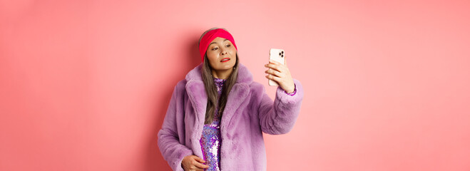 Fashion concept. Stylish asian senior female taking selfie on smartphone, posing in purple faux fur...