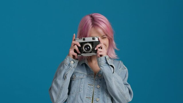 Isolated caucasian female photographer standing on blue background holding analogue camera. Hipster girl pretending to take photo of somebody. Wearing a denim jacket, looking directly at camera 4K