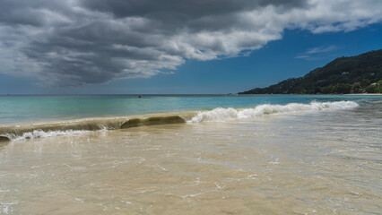 A long wave rolls onto the shore, twisting. Turquoise water mixes with the sand of the beach. Picturesque clouds in the blue sky. A hill in the distance. Seychelles. Mahe.Beau Vallon