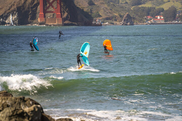 Men ride a hydrofoil surfboard using a hand held foil wing. 