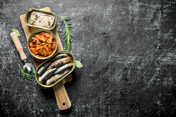 Open tin cans of canned fish on a cutting Board with a opener and rosemary.