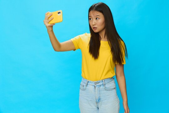 Asian woman holding her phone and looking at the screen taking pictures of herself talking on a video call with a yellow case on a blue background wearing a yellow T-shirt smiling with teeth 