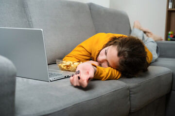 Teen girl watch bored movie on laptop while relax in the living room. She is lying on couch, eating apple chips, watch film at home.