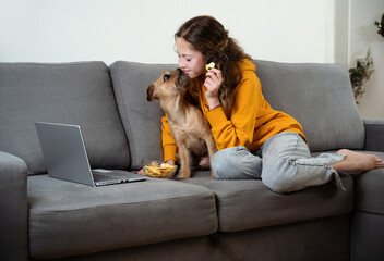 Teen girl watch movie on laptop while relaxing in the living room. She is sitting on couch, eating apple chips, watching video at home. Small dog pets wants snack.