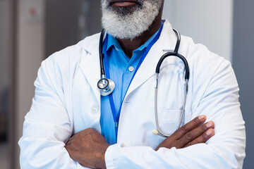 Midsection of african american senior male doctor with arms crossed in hospital corridor, copy space