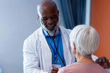 Fototapeta premium Happy diverse senior male doctor using stethoscope on senior female patient, copy space