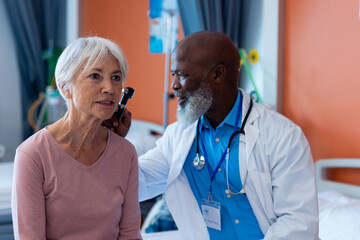Diverse senior male doctor using otoscope examining ear of senior female patient, copy space