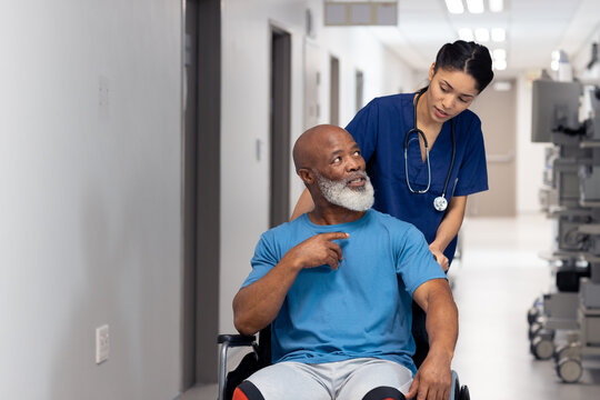 Diverse female doctor pushing senior male patient in wheelchair in corridor, copy space