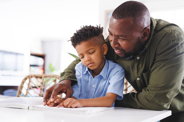 Happy african american father and blind son at table, reading braille